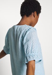 Jasnoniebieska bluzka z krótkim rękawem z haftowanymi detalami typu eyelet i luźnym krojem. Okrągły dekolt i marszczone rękawy nadają jej struktury.