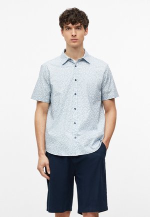 Jonge man met krullend haar die een lichtblauw, patroon overhemd met korte mouwen en donkerblauwe korte broek draagt, staand met één hand in de zak.