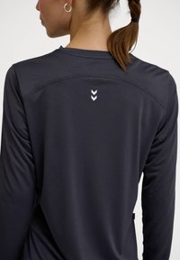 Długi rękaw sportowa bluzka w ciemnoszarym kolorze, wykonana z teksturowanej tkaniny, z okrągłym dekoltem i małym białym logotypem w kształcie chevronu na plecach.