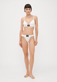 White lingerie set featuring a triangular bra with narrow straps and a matching thong. Both pieces have black branding accents.