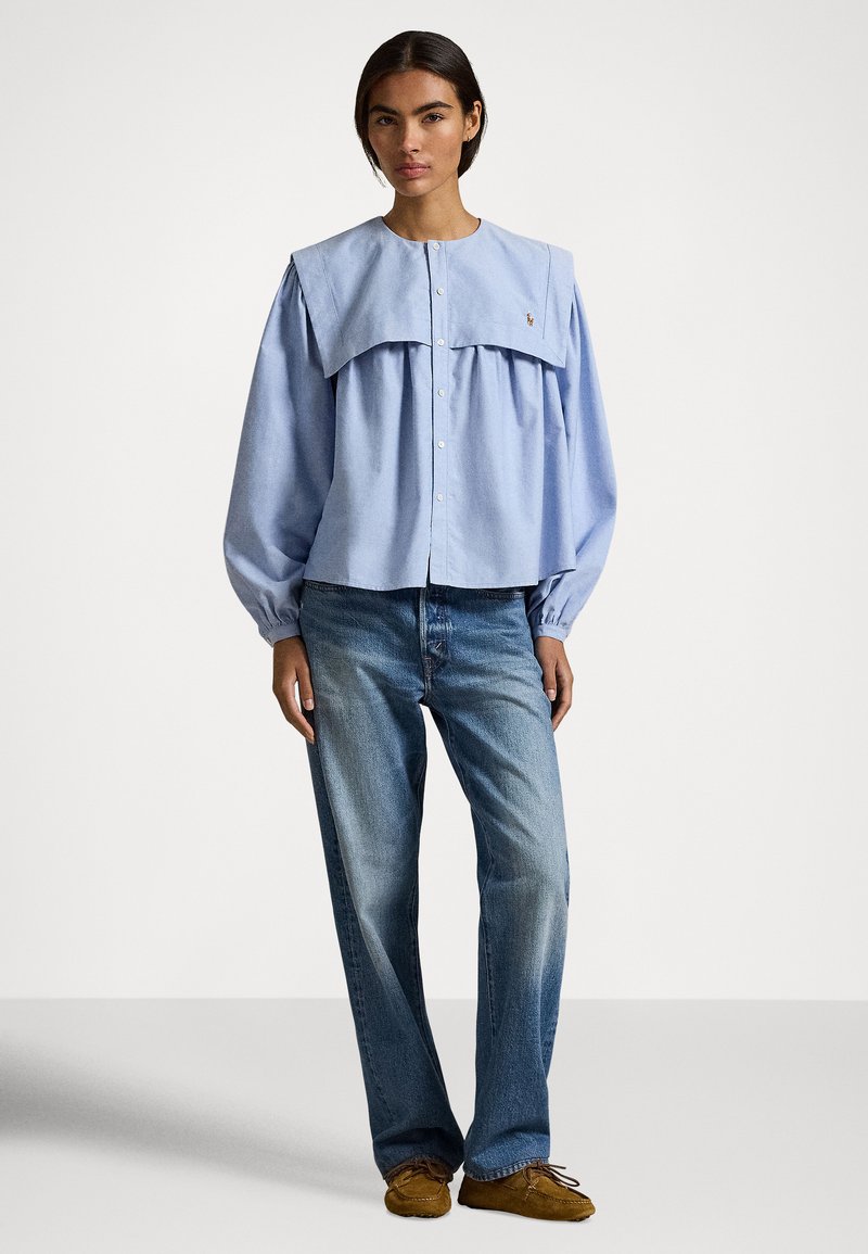 Vrouw draagt een lichtblauw blouse met een brede kraag, losse blauwe jeans en bruine mocassinschoenen, staand tegen een effen witte achtergrond.