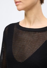 Κοντινό πλάνο ενός ατόμου που φοράει ένα μαύρο, χαλαρά πλεκτό τοπ από δίχτυ, αποκαλύπτοντας το δέρμα από κάτω, με στρογγυλή λαιμόκοψη.