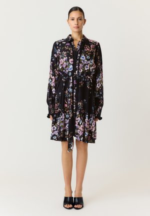 Femme portant une robe noire à fleurs à manches longues avec une ceinture à la taille et des chaussures à talons hauts ouvertes noires, debout devant un fond uni.