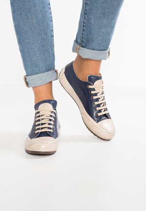 Pies con vaqueros remangados azules y zapatillas con cordones azul marino y beige sobre un fondo blanco.