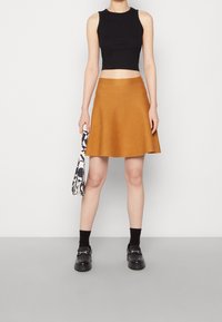 Top court noir, jupe évasée moutarde, chaussettes noires, chaussures noires imposantes avec quincaillerie, tenant un sac fourre-tout à motifs.