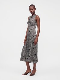 Femme debout portant une robe midi noire sans manches avec un motif floral blanc et des sandales noires à talons ouverts, sur un fond uni.