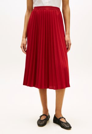 Kvinne iført et knelangt rødt plissert skjørt, hvit skjorte og svarte vevde Mary Jane-sko som står foran en ensfarget bakgrunn.