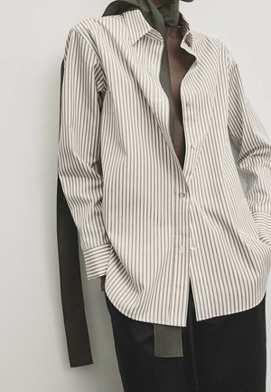 Personne portant une chemise blanche ample avec de fines rayures verticales noires, associée à un pantalon noir et une écharpe sombre autour du cou.