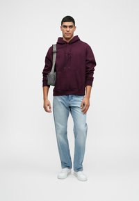 Sudadera con capucha en color burdeos y bolsillo frontal, combinada con jeans azul claro y zapatillas blancas. Pequeño bolso cruzado en gris. Estilo casual.