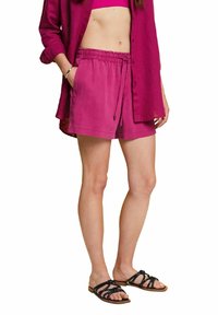 Femme portant un short magenta à cordon, une chemise ouverte assortie, et des sandales plates noires à lanières, debout avec une main dans la poche.