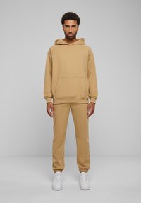 Beige hettegenser med kengurulomme, matchende tapered joggebukser og hvite sneakers. Myk tekstur og uformell design.
