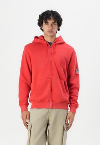 Crvena zip-up dukserica s kengurufrontom i kapuljačom na šnuru. Ima suptilni logo i oznaku američke zastave na lijevom rukavu.