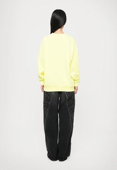 Neon gul oversized sweatshirt med ribbede manchetter, parret med sorte vidde jeans med et tekstureret geometrisk mønster på siderne.