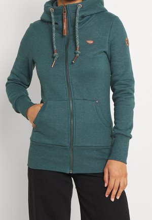 Persoon die een tealkleurige hoodie met ritssluiting, koordjes en voorzakken draagt, gecombineerd met een zwarte broek, staand tegen een effen achtergrond.