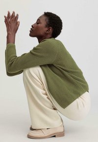 Grüner Strickpullover mit gerippter Textur, cremefarbene weite Hose und beigefarbene Slipper. Model posiert hockend und präsentiert die Lagen und Passform des Outfits.