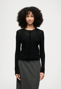 Ženska s kodrastimi lasmi, oblečena v črno kardigan z gumbi in temno siva krila, stoji pred enostavno svetlo ozadje.