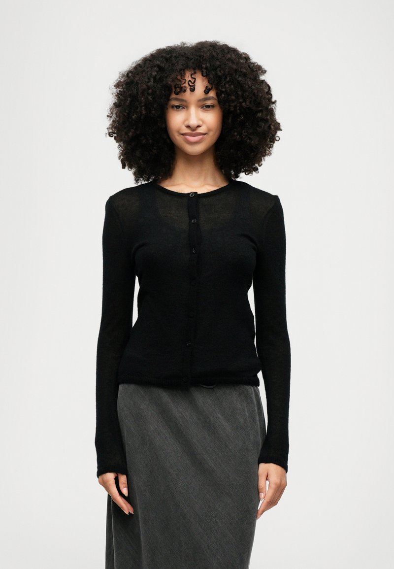 Ženska s kodrastimi lasmi, oblečena v črno kardigan z gumbi in temno siva krila, stoji pred enostavno svetlo ozadje.