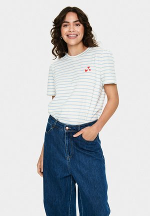 T-shirt rayé blanc et bleu clair à manches courtes, avec trois accents en forme de cœur rouge sur le côté gauche de la poitrine, associé à un pantalon en denim bleu.