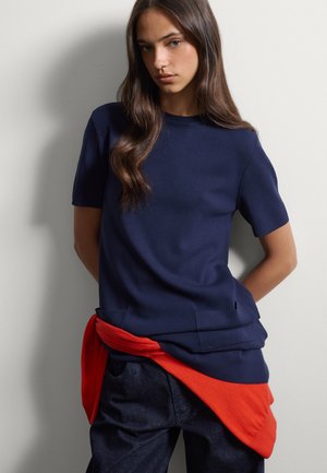 Mujer con cabello largo y castaño lleva una camiseta azul marino de manga corta, prenda roja atada a la cintura y pantalones oscuros, de pie contra una pared blanca.