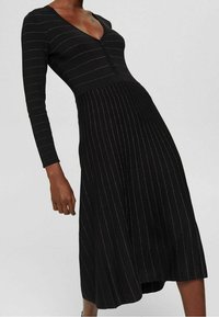 Femme portant une robe midi noire à manches longues avec de fines rayures verticales et horizontales et un décolleté en V profond avec des boutons.