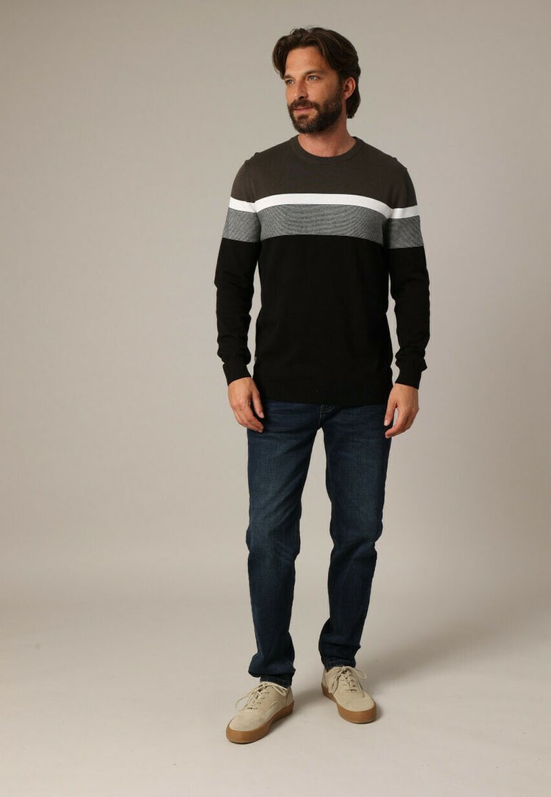 Pull rayé noir et gris à manches longues, en tissu texturé, associé à un jean bleu et des baskets beiges, se tenant sur un fond neutre.