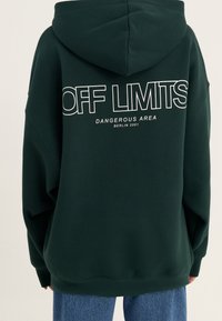 Sudadera verde con un gran diseño de texto blanco en la parte posterior: "ÁREA PELIGROSA SIN ACCESO BERLÍN 2001." Corte holgado y tejido suave.