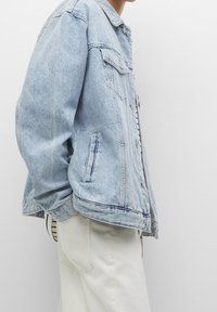 Personne portant une veste en jean bleu clair et un pantalon blanc cassé, debout de profil sur un fond gris clair uni.