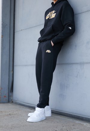 Person iført sort Nike Air træningsdragt og hvide høje sneakers, stående med hænderne i lommerne mod en grå væg.