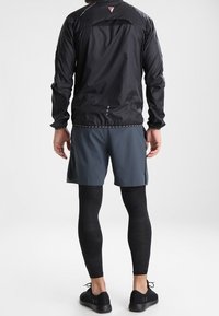 Veste noire légère avec dos en mesh, shorts gris et leggings noirs texturés. Des chaussures de sport noires complètent le look de sport.