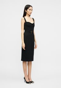 Γυναίκα με μακριά σκουρόχρωμα μαλλιά που φοράει στενή μαύρη midi φόρεμα χωρίς μανίκια με διακοσμητική ζώνη και μαύρες μυτερές γόβες.