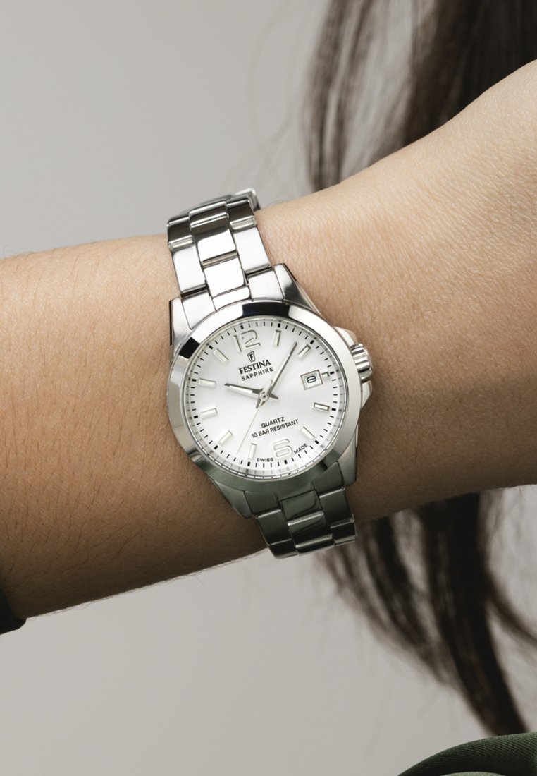 Reloj de cuarzo de acero inoxidable plateado con una esfera blanca, ventana de fecha y pulsera de eslabones. Las características notables incluyen un cristal de zafiro.