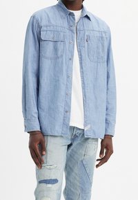 Camicia di jeans azzurro chiaro con maniche lunghe, frontale con bottoni e due tasche sul petto. Indossata sopra una t-shirt bianca e jeans azzurro chiaro a patchwork.