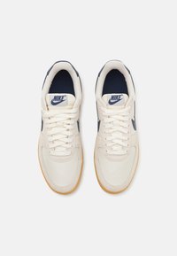Nike sporta apavi ar krēmkrāsas auduma virsu, tumši zila akcentiem un gumijas zoli. Iekļauj plakanas šņores un logotipu uz mēles. Plakana, apaļa purngala forma.