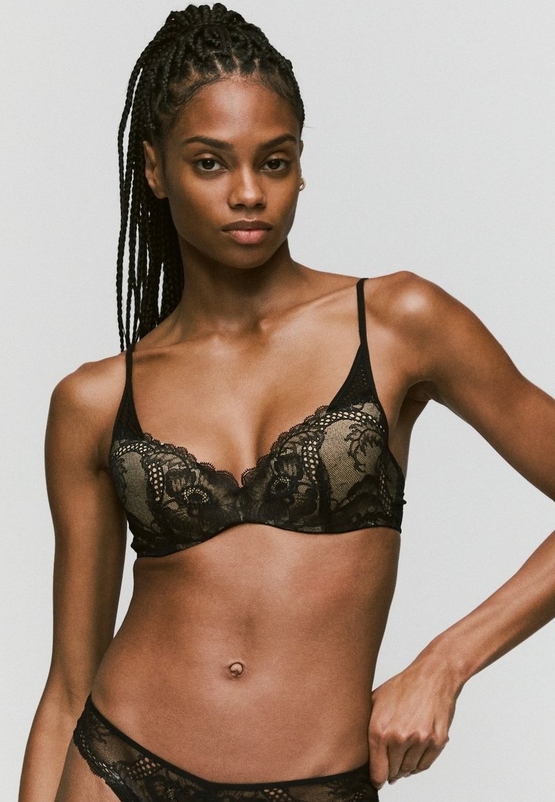 Sujetador de encaje negro con patrones florales, tirantes ajustables y forma triangular, que enfatiza un diseño intrincado y una textura transparente.