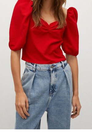 Kobieta w jasnoczerwonym topie z bufkami połączonym z jasnoniebieskimi plisowanymi dżinsami z wysokim stanem na jednolitym tle.