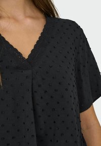 Black short-sleeve top featuring a V-neckline, textured fabric with raised dots, and delicate lace trim along the neckline.