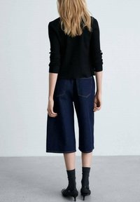 Culottes en denim noir avec jambes larges, associées à un pull noir ajusté. Cheveux longs et ondulés visibles. Chaussures noires à talons bas portées avec des chaussettes grises.