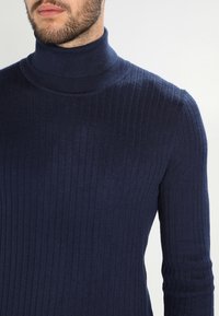 Marinblå ribbad polotröja i mjukt material, med en figursydd design och texturerat mönster längs ärmarna och kroppen.