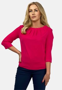 Fuksjowa bluzka z plisowanym dekoltem i rękawami trzy czwarte, uszyta z gładkiego materiału, w parze z granatowymi spodniami.