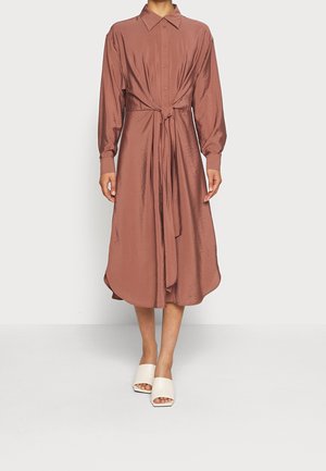 Kvinde iført midi-kjole med lange ærmer og knapper, bindebånd i taljen i skinnende rustfarvet stof, kombineret med hvide sandaler med åben tå.