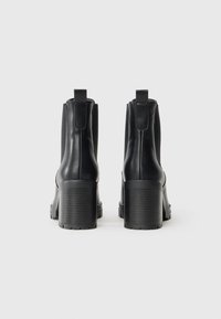 Mustad musta värvi ankle'ite saapad, millel on sile nahkviimistlus, kummist tald ja paks konts. Omab külgmisi elastseid paneele ja tõmblinti taga.