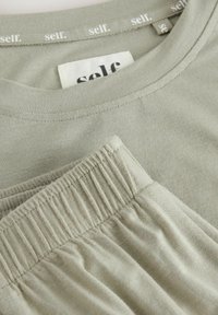 Beige bomulds T-shirt og bukser med elastisk talje, hvor "self." mærket er synligt på kraven og indersiden af etiketten.