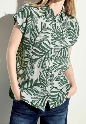 Femme portant une chemise à manches courtes boutonnée avec un imprimé de feuilles vertes et un jean bleu foncé, debout les mains dans les poches.