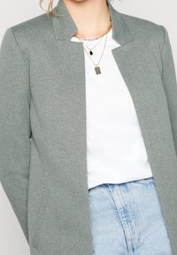 Blazer gris clair avec col cranté, en tissu texturé. Porté sur un haut blanc, associé à un jean bleu clair. Colliers dorés superposés.