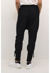 Pantalons de jogging noirs avec une taille élastique, des ourlets resserrés et des cordons de serrage sur le côté, fabriqués à partir d'un tissu léger, associés à des baskets blanches.