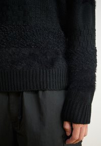 Black knitted sweater featuring varying textures, including smooth and fuzzy sections, with a ribbed hem and cuffs. Worn with black pants.