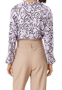 Lila blus med ett svart blommönster, med lös passform och långa ärmar, i kombination med beiga skräddarsydda byxor.