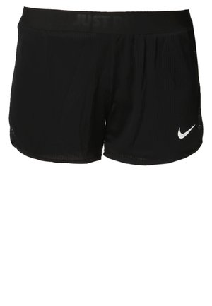 Kvinde iført hvid V-hals T-shirt, sorte Nike-shorts og sorte Nike-sneakers, gående fremad mod en ensfarvet hvid baggrund.
