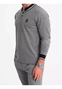 Polo gris à manches longues avec un col noir orné de rayures blanches, trois boutons et un logo brodé sur la poitrine.