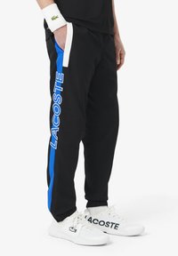 Zwarte joggingbroeken met blauwe zijkanten en "LACOSTE" merknaam. Gemaakt van zacht materiaal met een elastische tailleband en boorden bij de enkels.